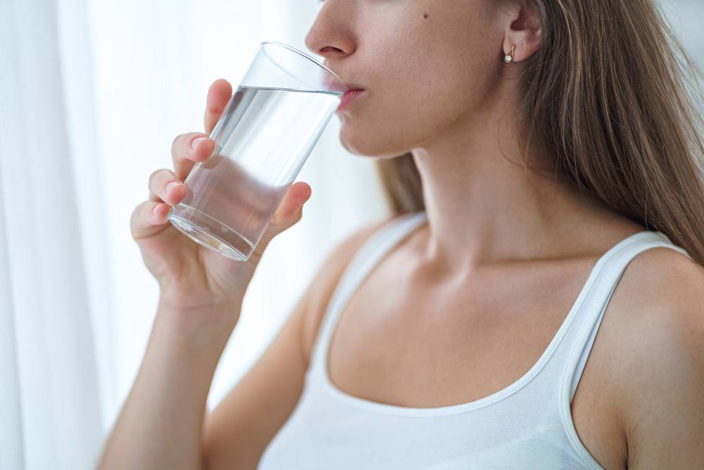 Mulher bebendo um copo de água cristalina, com fundo claro e ambiente calmo, simbolizando a importância de beber água para a saúde e o bem-estar.