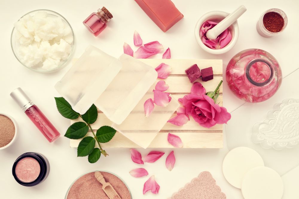 Vista superior de ingredientes para cosméticos artesanais, incluindo base para sabonete, creme hidratante caseiro, óleo de coco, óleo essencial, água com infusão de rosas e pó de argila rosa.